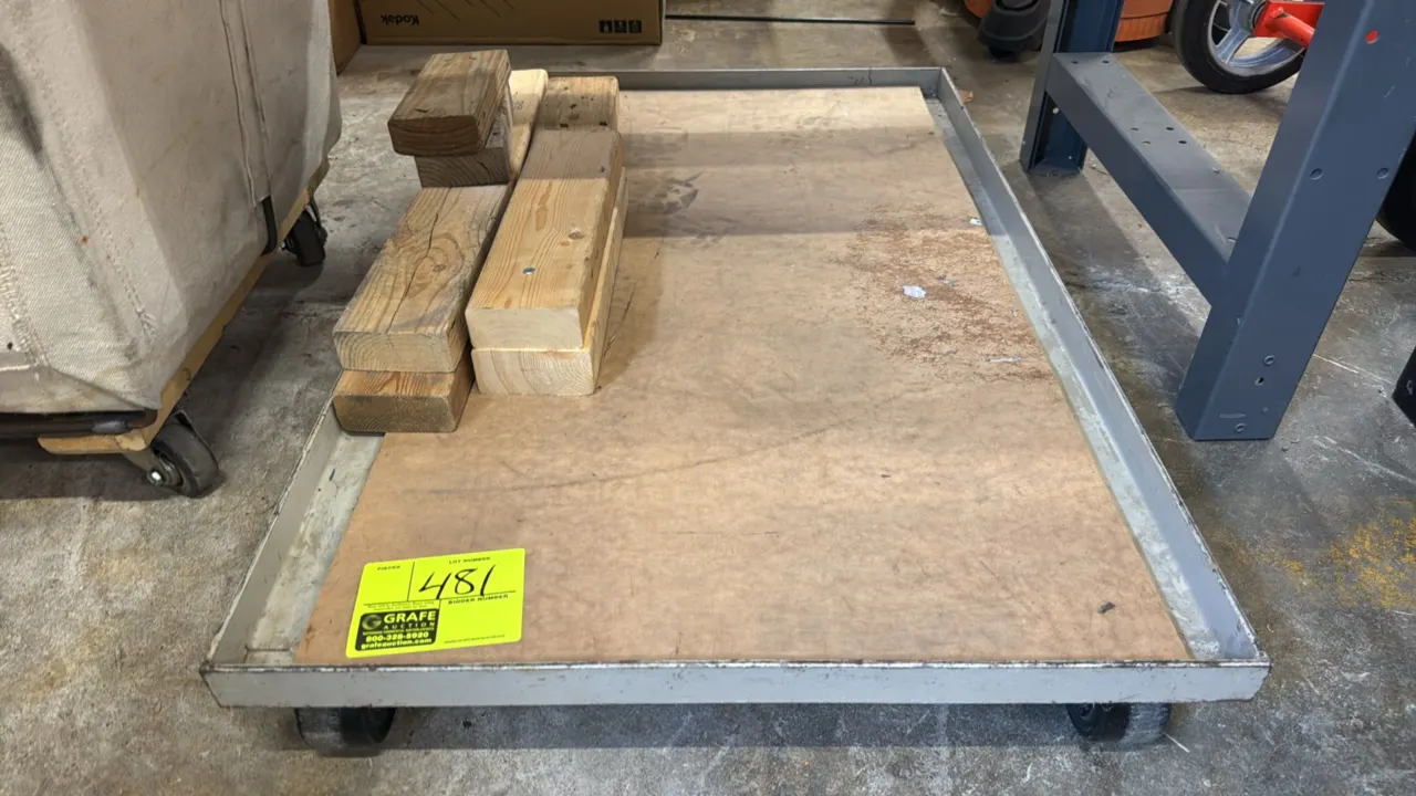 Metal 4-wheel platform dolly. Features a raised-lip metal frame with a wood deck insert. Includes wood blocks shown. 24" x 37". Mounted on 4 casters.