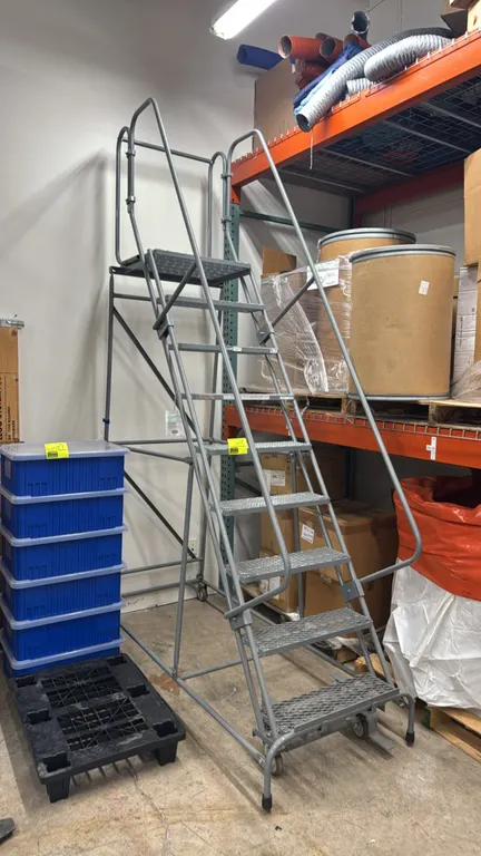 Uline 9-step portable steel stock ladder. Features expanded metal grating treads, top platform, safety handrails, and a lockstep mechanism. Rated for