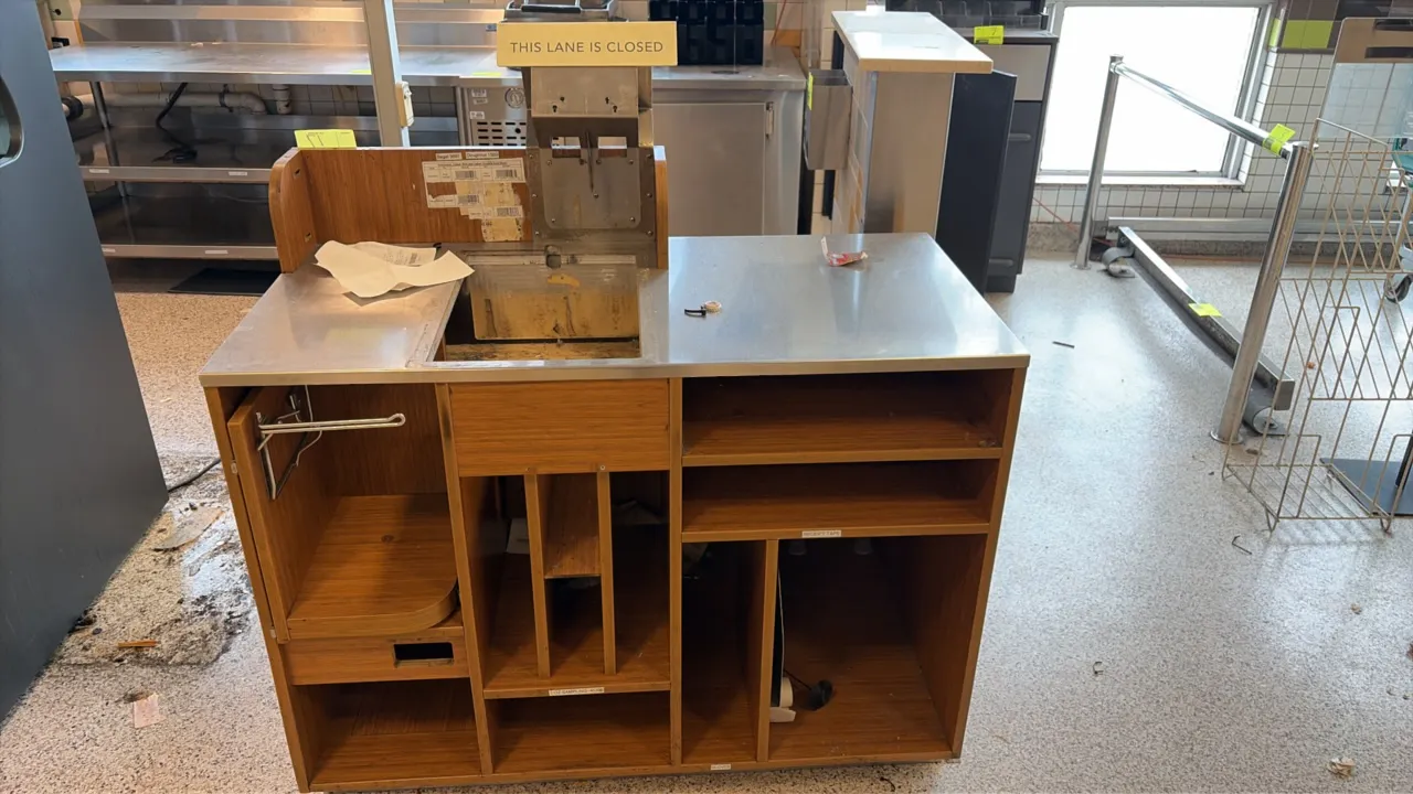 Checkout lane counter. Features a stainless steel countertop with scanner/scale cutout, wood-grain laminate exterior, cashier-side storage shelving an