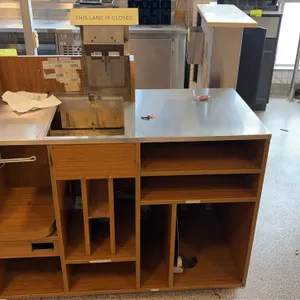 Checkout lane counter. Features a stainless steel countertop with scanner/scale cutout, wood-grain laminate exterior, cashier-side storage shelving an