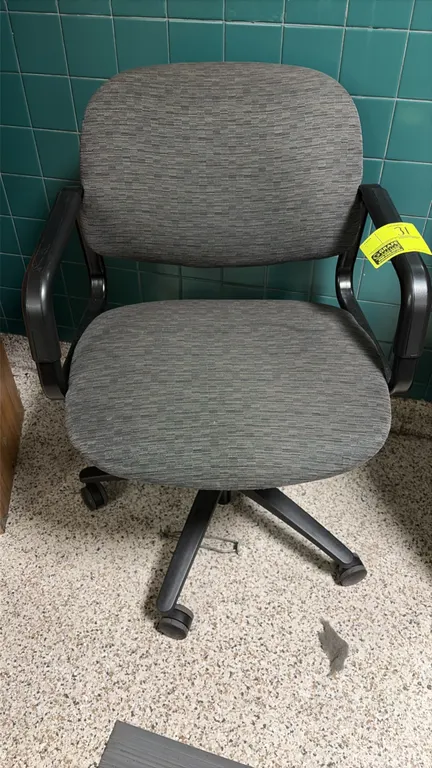 Office chair. Features gray textured fabric upholstery, padded seat and backrest, and fixed black armrests. Mounted on a 5-star swivel base with caste