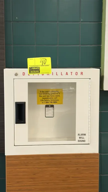 Metal AED defibrillator storage cabinet. Features white finish, hinged door with viewing window, recessed pull handle, and key lock hole. Wall-mount d