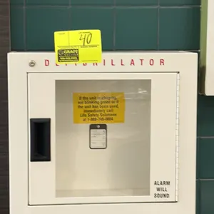 Metal AED defibrillator storage cabinet. Features white finish, hinged door with viewing window, recessed pull handle, and key lock hole. Wall-mount d