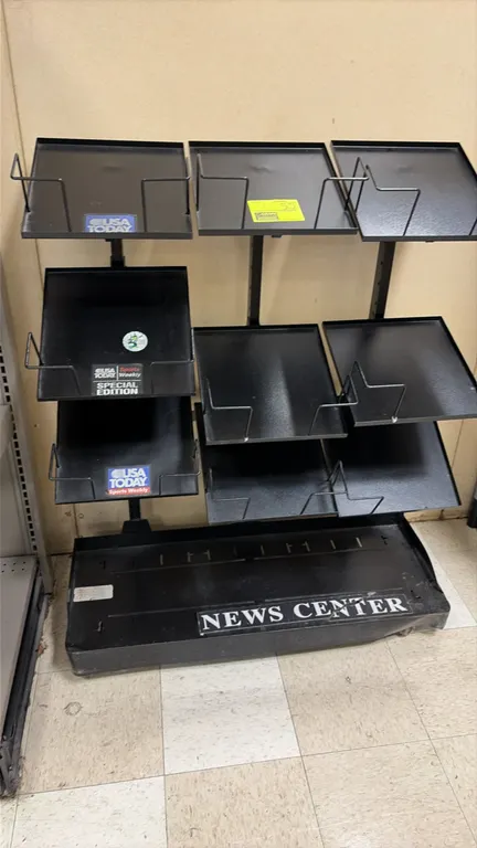 Metal portable newspaper display rack. Features multi-tier angled metal shelves with wire retainers, lower base shelf with "NEWS CENTER" labeling, and