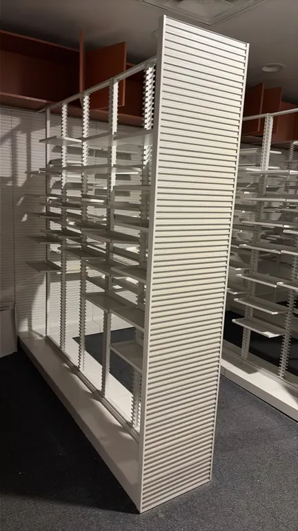Double-sided pharmacy shelving unit. Features slotted uprights, adjustable metal shelves, base deck, and slatted end panels. Finished in white. Backgr