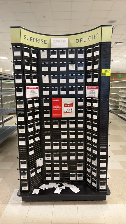 Retail gift card display rack. Features multi-sided black metal pegboard panels, display hooks, and an upper signage canopy header with 'Surprise Give