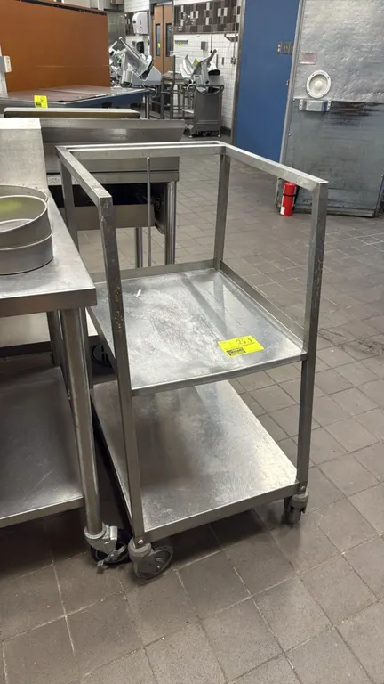 Stainless steel 2-tier utility cart. Features stainless steel construction, two solid shelves, and an open upper frame. Mounted on swivel casters.
