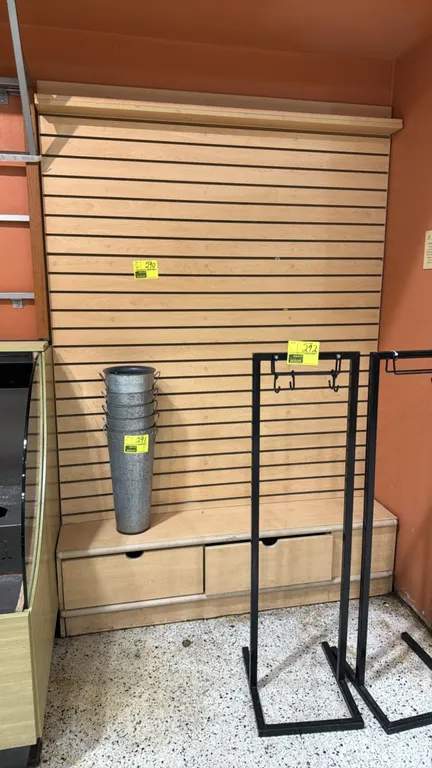 Slatwall display fixture with base cabinet. Features a light woodgrain laminate finish, horizontal slatwall panel, upper header shelf, and bottom stor