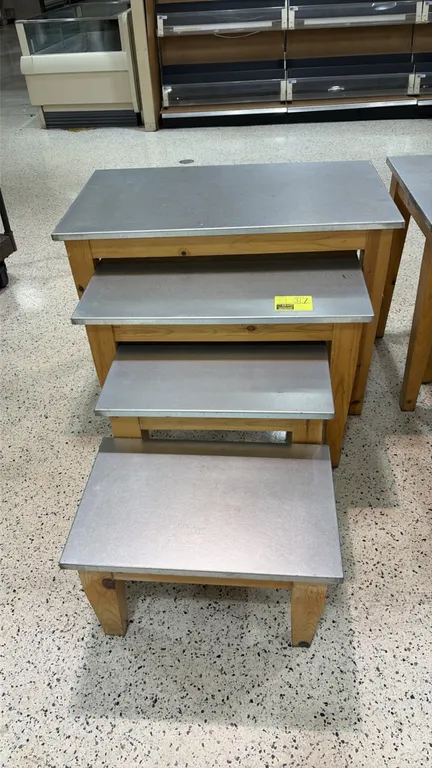 Set of (4) assorted sized nesting display tables. Features sheet metal tops over natural wood bases and legs. Graduated sizing allows for compact nest
