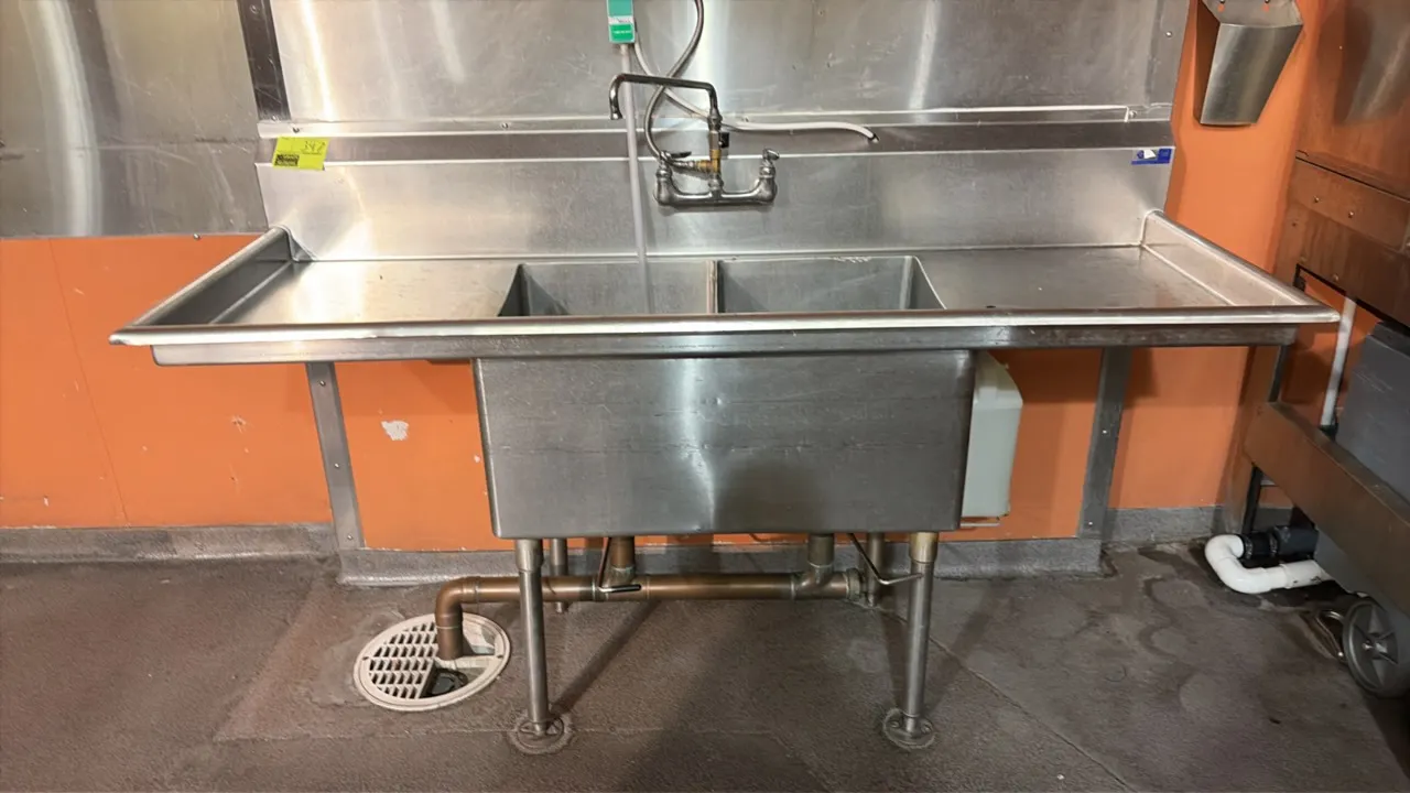 Stainless steel 2-compartment sink. Features left and right drainboards, backsplash, and splash-mounted faucet. 72" x 26 1/2". Mounted on tubular legs