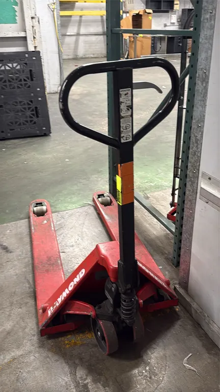 Raymond manual pallet jack. Features steel forks, hydraulic lift pump handle with fingertip release lever, dual steer wheels, and nylon load rollers.