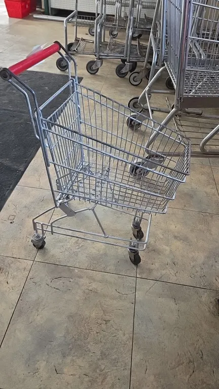 Kid-size metal shopping cart. Features wire basket construction and a red plastic handle grip. Mounted on (4) casters.