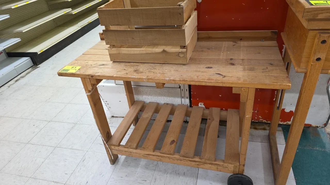 Portable wood display table with wood crate. Features a slatted wood top with raised border rail, lower slatted undershelf, and two wheels for tilt-an