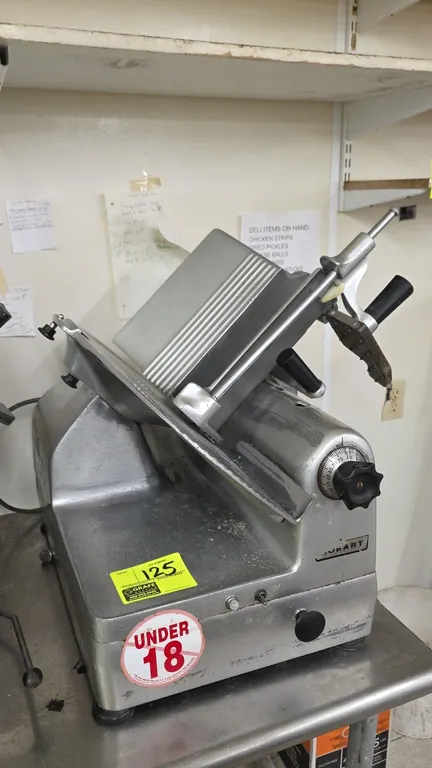 Hobart commercial deli slicer. Features an angled gravity-feed product tray, adjustable slice thickness indexing knob, weighted product pusher, and to