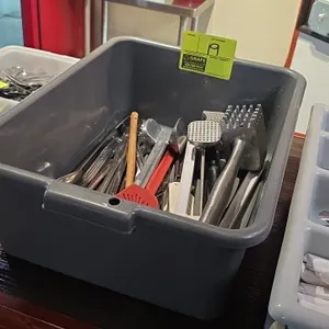 Assorted kitchen smallwares with gray plastic bus tub. Includes meat tenderizer mallets, spatulas, slotted spoons, ladles, and dial thermometer.