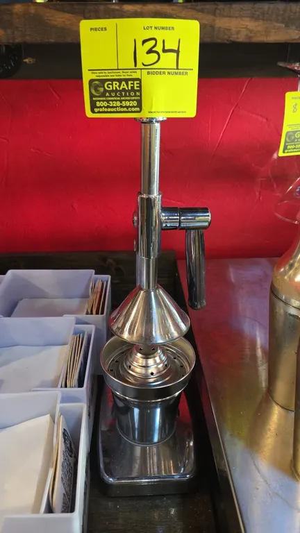 Stainless steel manual citrus juicer. Features manual lever-action press handle, perforated strainer cone, and collection cup. Countertop base.