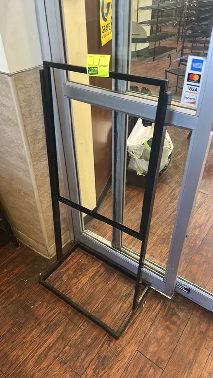 Metal sign stand. Features a black metal frame designed for slide-in sign inserts. 2'W. Mounted on a rectangular metal floor base.