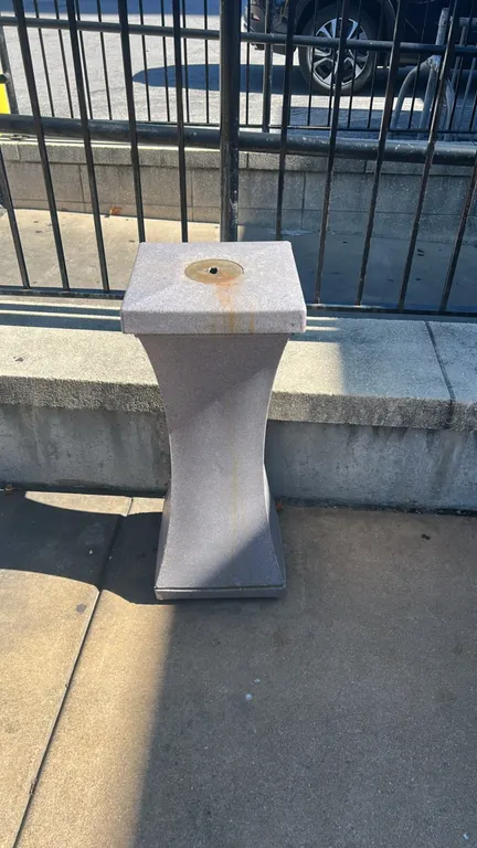 Polyethylene outdoor cigarette receptacle. Features hourglass pedestal design, textured faux-stone finish, and top metal snuff plate.