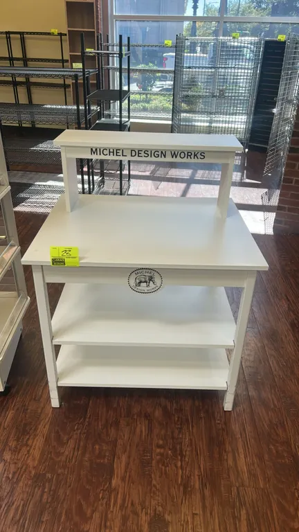 Michel Design Works 3-tier laminate merchandising table. Features a white finish, top riser shelf, two lower shelves, and Michel Design Works branding