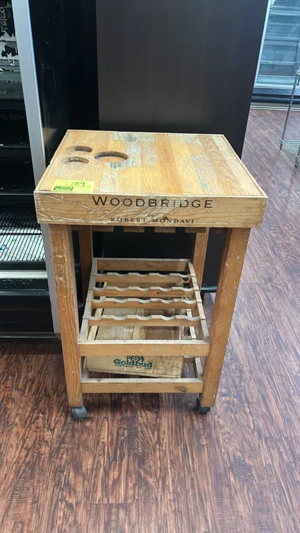Woodbridge by Robert Mondavi wooden mobile wine cart. Features top surface bottle cutouts, under-mount stemware rack, lower wine bottle rack, and bott