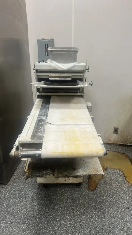 Commercial portable bakery dough sheeter and moulder. Features conveyor belt table, adjustable roller controls, metal infeed chute, and lower catch tr