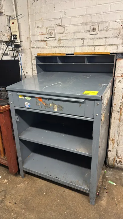 Metal shop receiving desk. Features upper organizer compartments, single pull-out drawer, and two lower open storage shelves. Gray metal construction.