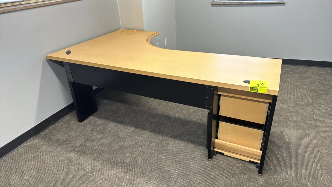 Wood laminate corner desk. Features light woodgrain laminate top, black frame, two built-in storage drawers with bar pulls, and desktop cable grommets