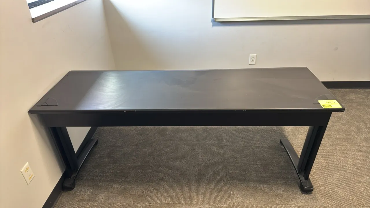 Laminate office table. Features a black laminate work surface with dual corner cable grommets. Mounted on a black metal base with T-style legs. Dimens