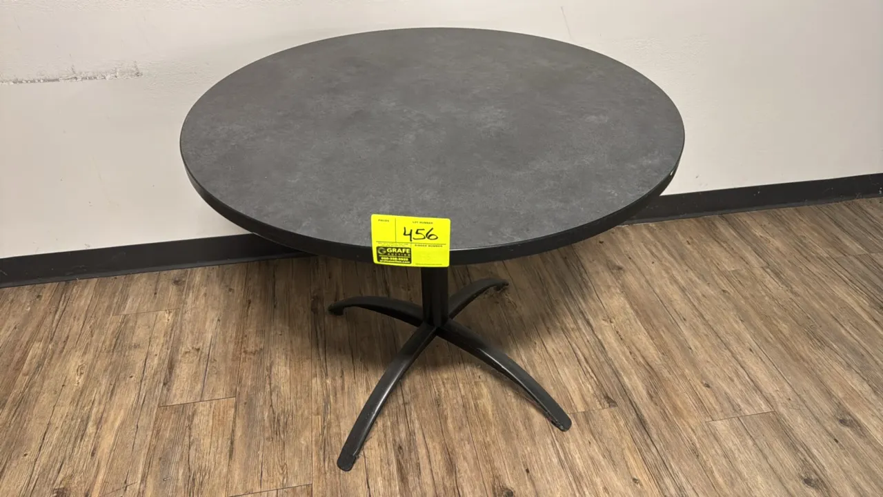 Round laminate table. Features a dark slate-look laminate top with black protective edge banding. 3' Dia. Mounted on a black metal pedestal base with