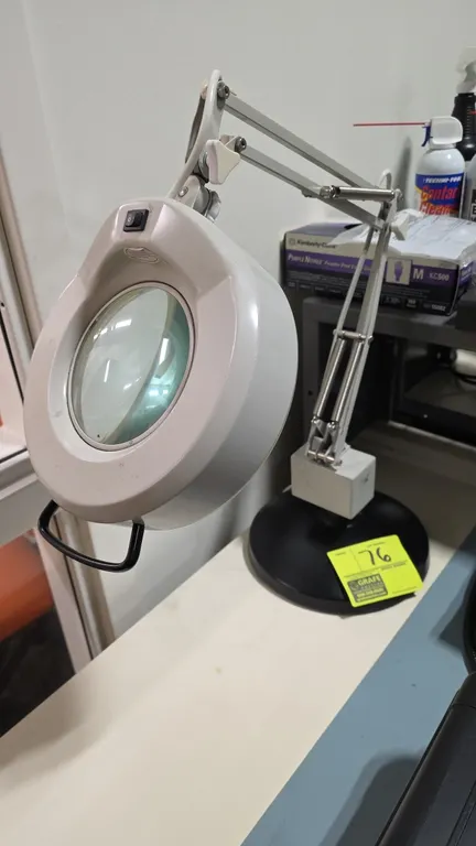 Adjustable magnifying task light. Features an articulated spring-balanced swing arm, built-in magnifying lens, circular lamp housing, top handle, and