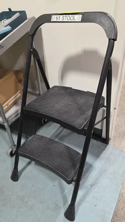 2-step folding step stool. Features a black tubular metal frame, textured slip-resistant plastic steps, upper molded handle grip, and rubber feet.