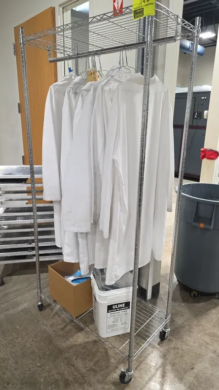 Chrome mobile wire garment rack. Features top and bottom wire shelves and a hanging bar. 36"W x 18"D x 77"H. Mounted on swivel casters. Contents not i