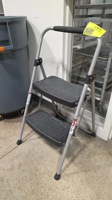 2-step folding step stool. Features a tubular metal frame, two textured molded plastic steps, and upper padded hand grip.