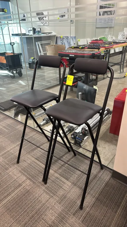 Metal folding bar stools. Features black vinyl padded seats and backrests, folding black tubular metal frames, and integrated footrests.