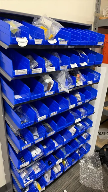 Multi-tier parts rack with blue plastic storage bins. Includes assorted contents such as parts, gaskets, tubing, and fuses. Features multi-shelf metal
