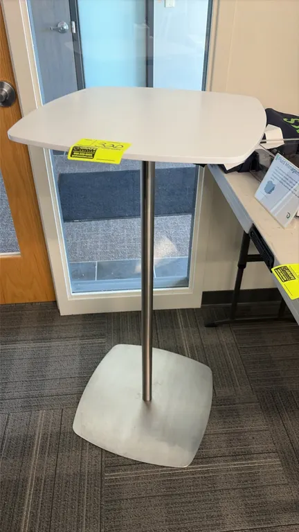 High-top table. Features a white square laminate top with rounded corners. 21" x 21". Mounted on a brushed metal pedestal base.