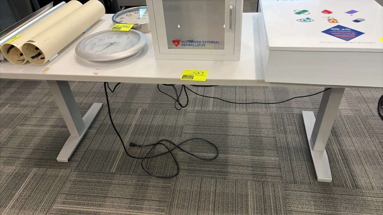 Electric height-adjustable work table. Features a wood laminate top and digital push-button control panel with programmable presets. Mounted on a gray