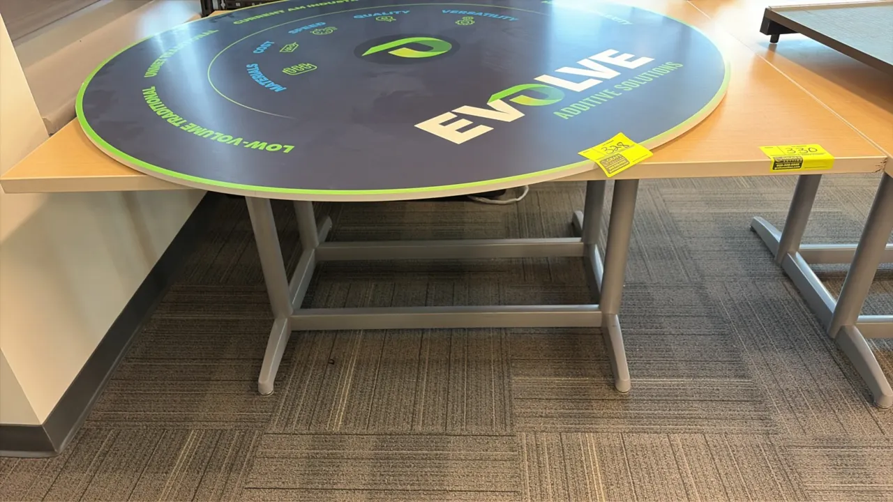 Wood laminate workstation. Features an integrated electric plug-in. 60" x 48". Mounted on a gray metal base. Sign on top not included.