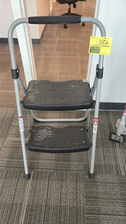 2-step folding step stool. Features a tubular metal frame, textured non-slip plastic treads, padded top hand grip, and rubber feet.