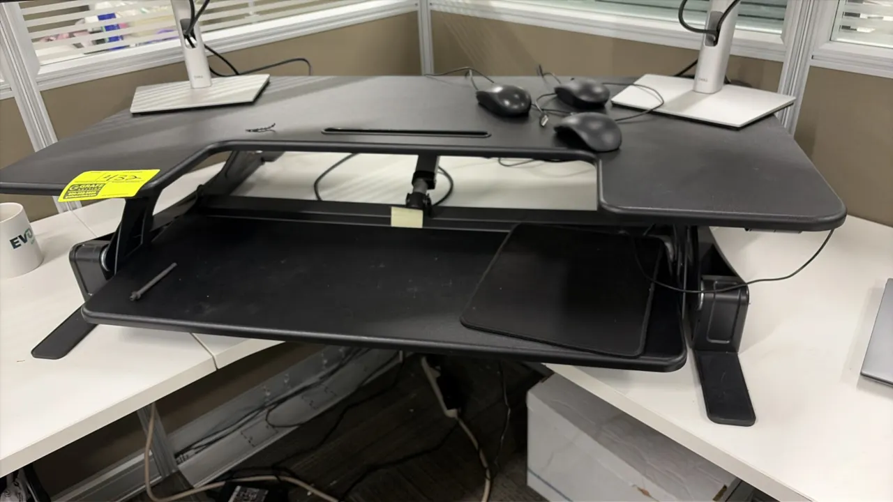 Electric sit-stand desktop riser converter. Features a two-tier design with an upper work surface, integrated device slot, lower keyboard tray, and bl