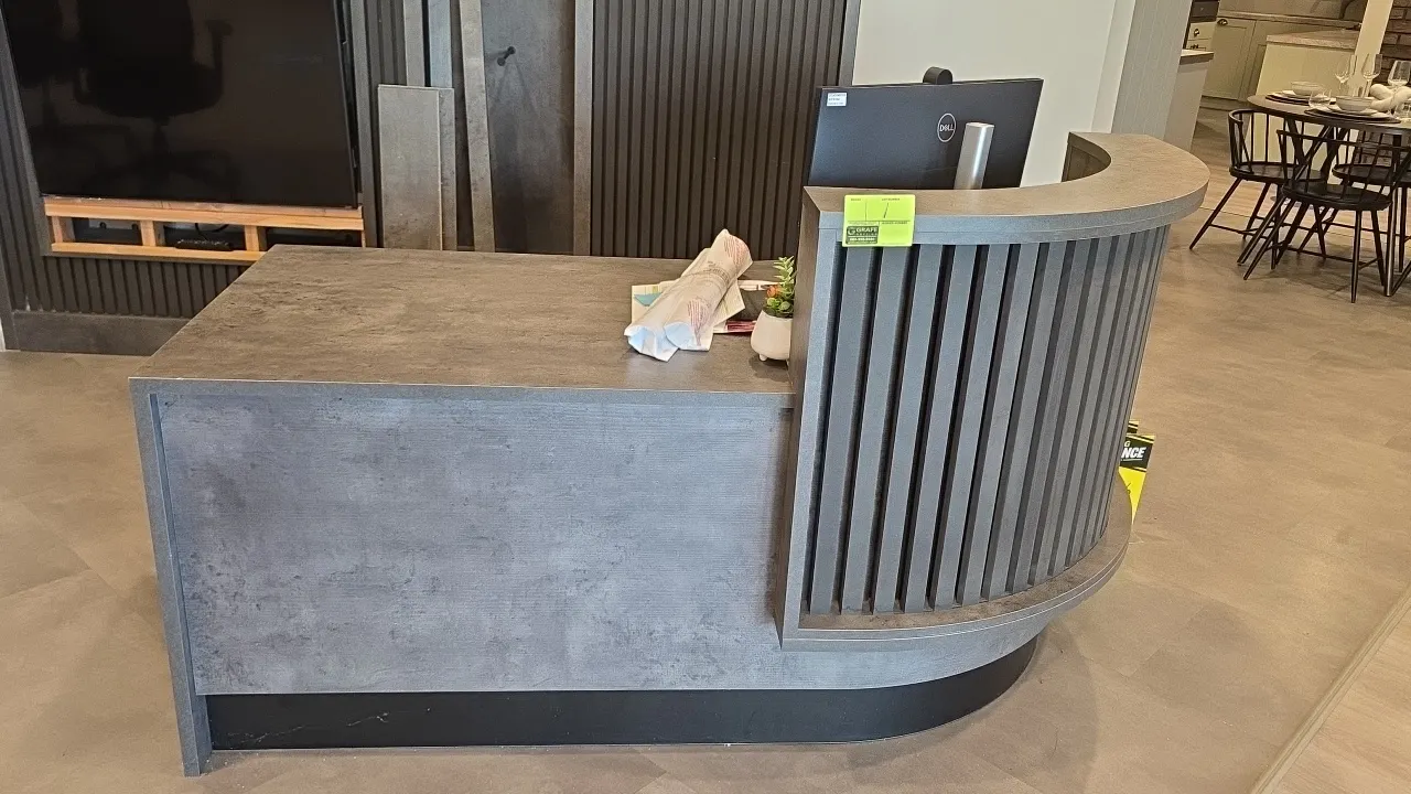 Laminate L-shaped reception desk. Features a curved transaction counter with vertical slatted accenting, textured concrete-look finish, single locking