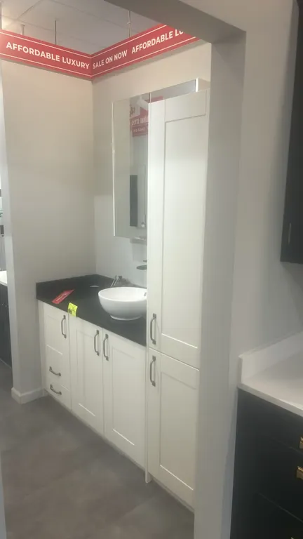 Infinity Plus bathroom vanity cabinet set. Features white laminate shaker-style base cabinets, matching tall linen cabinet, dark solid surface counter