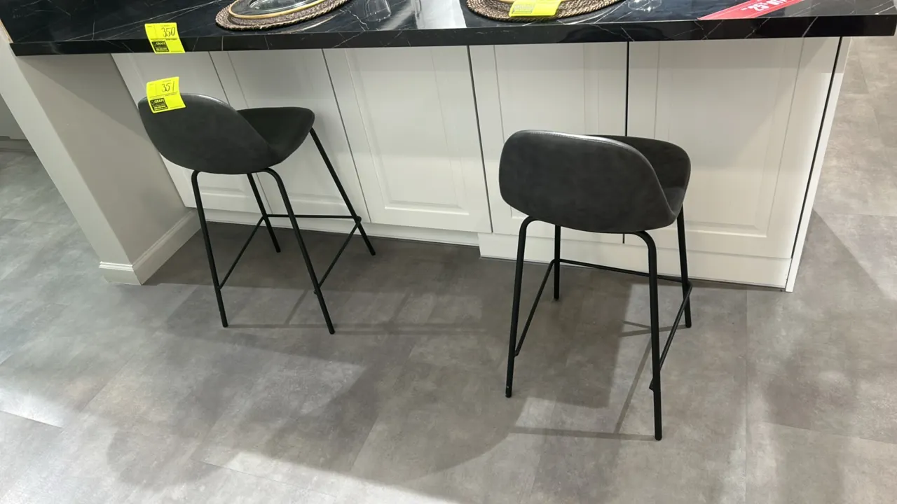 Cushioned barstools. Features charcoal gray faux leather cushioned seats with accent stitching. Mounted on black metal frames with integrated footrest