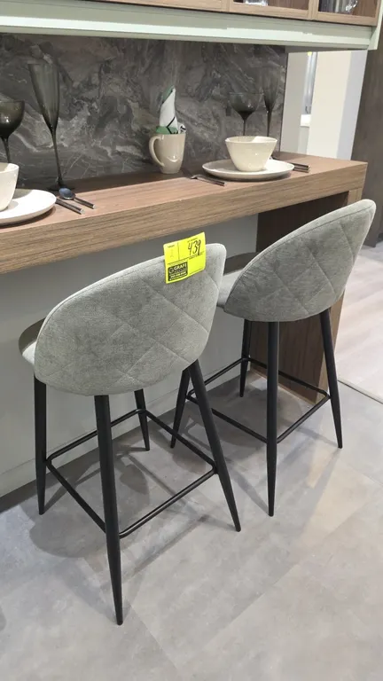 Upholstered barstools. Features gray fabric upholstery with diamond-quilted backrests, curved seats, and black metal legs with integrated footrests. 2