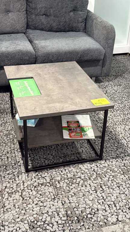 Laminate top coffee table. Features a gray weathered-finish laminate top, lower open storage shelf, and black metal frame. 31.5" x 23.5". Contents not