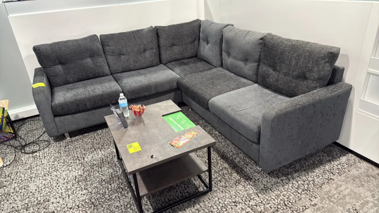 L-shaped sectional couch. Features gray textured fabric upholstery, tufted back cushions, and chrome-finish legs. 87" x 59". Coffee table and foregrou