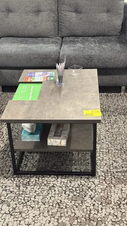 Laminate top coffee table. Features a weathered gray laminate top and lower storage shelf. 31.5" x 23.5". Supported by a black metal frame. Contents a