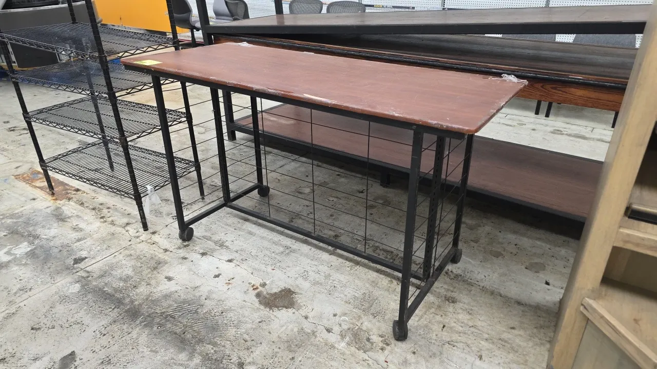 Mobile table cart with laminate wood top. Features black metal frame with wire grid sides and back panel. 64"L. Mounted on casters.