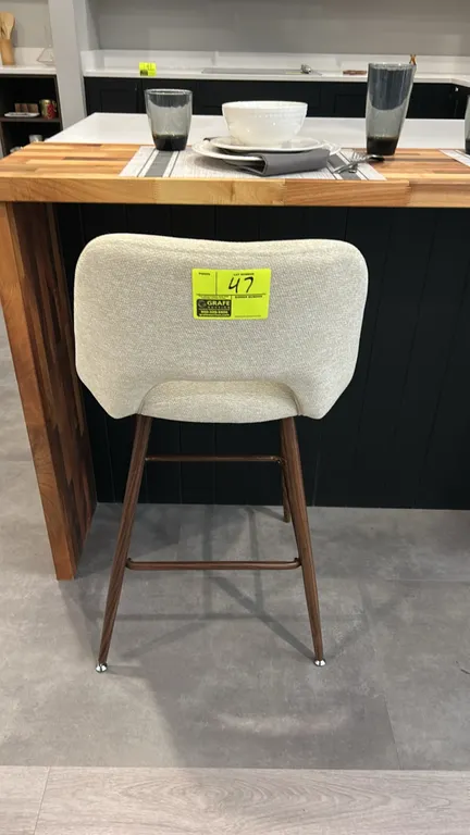 Cushioned barstool. Features textured light fabric upholstery with cutout back design, padded seat, tapered wood-finish legs, and integrated footrest