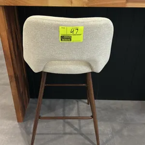 Cushioned barstool. Features textured light fabric upholstery with cutout back design, padded seat, tapered wood-finish legs, and integrated footrest 